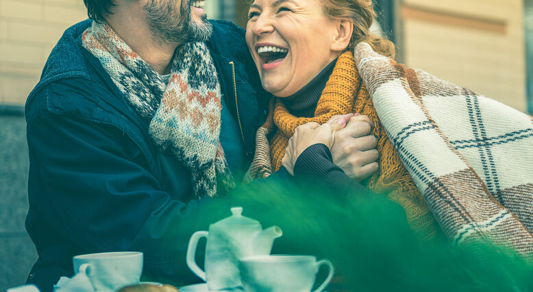 Ein Paar sitzt eingekuschelt im Freien beim Kaffeetrinken. Er umarmt sie, beide lachen.